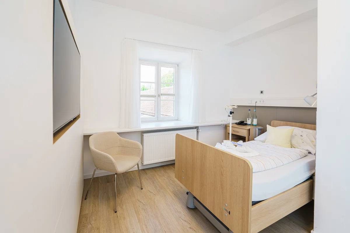 A bright patient room at St. George Hospital, with a wooden-framed bed and an armchair.