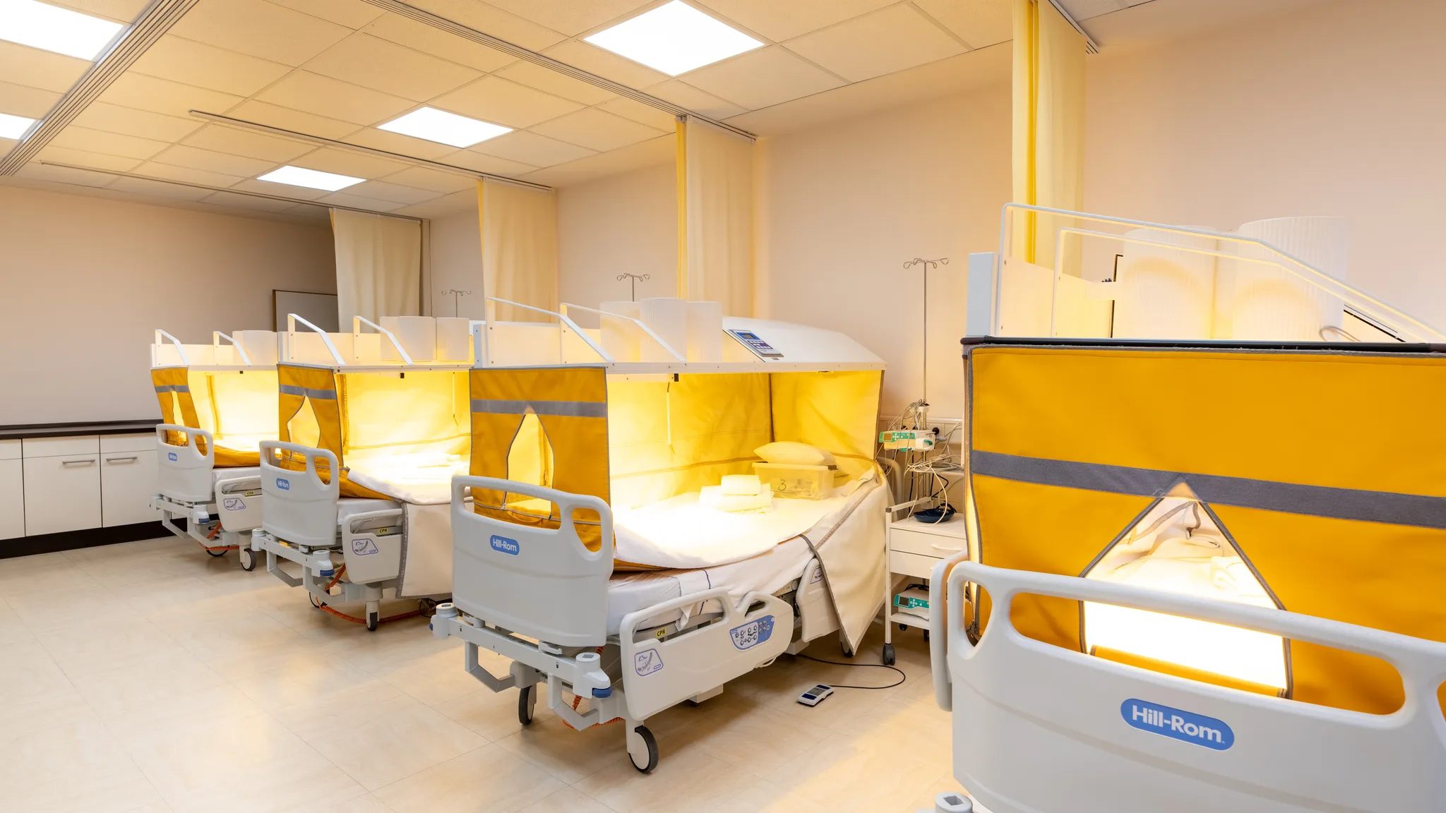 Four hospital beds under illuminated yellow infrared canopies in the whole-body hyperthermia room.