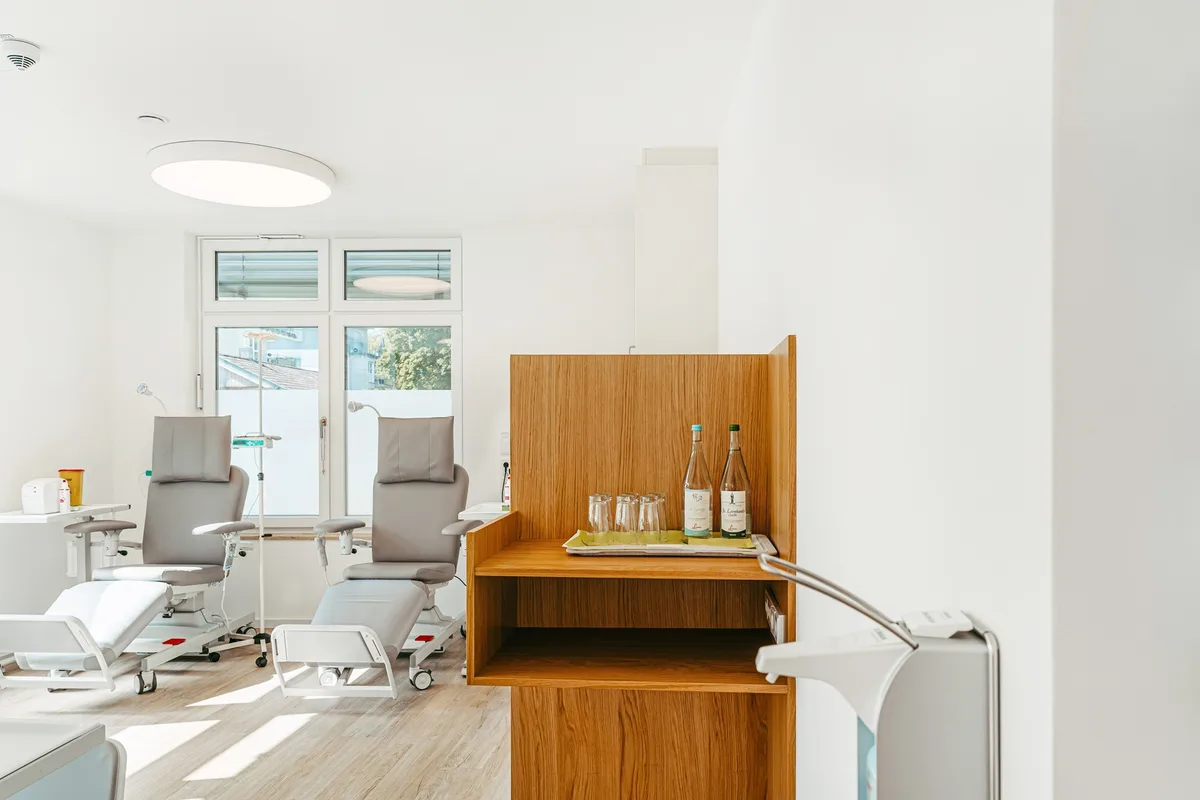 An infusion room with pale reclining chairs beside a wooden cabinet and a bright window